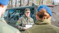 Eine Soldatin steht vor einem Fahrzeug und spricht zu einer Gruppe Soldatinnen und Soldaten 