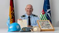 Ein Soldat sitzt am Tisch. Vor ihm liegen ein Helm, ein Barett und Fotos. Er hält eine Urkunde in der Hand.