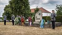 Auf einer Steinplatte steht ein Gedenkstein. Davor stehen bunte Kränze und eine Formation Soldaten.