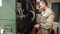 Ein Soldat steht an einem großen Serverschrank und hält diverse Kabel in den Händen