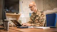 Ein Soldat sitzt am Schreibtisch und tippt auf der Tastatur seines Laptops