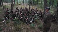 Eine Gruppe junger Menschen sitzt im Wald und hört einem Ausbilder zu.