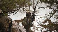 Ein Soldat steht mit seinem hochgehaltenen Gewehr auf einem Felsen im bewaldeten Gebirge.