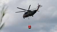 Ein Transporthubschrauber NH-90 aus der Froschperspektive, an dem ein großer Kanister für Löschwasser angehängt ist