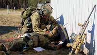 Hinter einer Deckung legt ein Soldat einem verwundeten Kameraden am Boden einen Verband an.