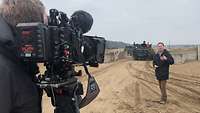 Ein Reporter steht mit Mikrofon vor einer Kamera, im Hintergrund zwei Schützenpanzer.
