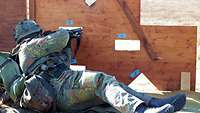 Ein Soldat liegt hinter einer hölzernen Wand mit Schießscharten und schießt mit seinem Gewehr. 