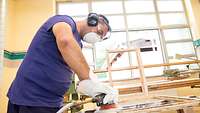 Ein Mann mit Schutzbrille und Handschuhen schleift einen Holz-Fensterrahmen