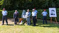 Drei Männer stehen vor einem Gedenkstein. Man sieht ein Porträt von Stauffenberg. 