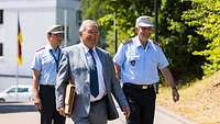 Drei Männer, zwei davon in Uniform, gehen zu einer Gedenkveranstaltung. Im Hintergrund weht eine Deutschlandfahne.