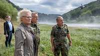 Christine Lambrecht steht mit weiteren Soldaten auf einer Wiese, im Hintergrund nimmt ein Hubschrauber Löschwasser auf.