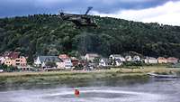 Löscharbeiten Sächsischen Schweiz Ein Mehrzweckhubschrauber vom Typ NH-90 befüllt den Löschwasser-Außenlastbehälter. Im Hintergrund stehen Häuser am Ufer.