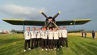 Eine Gruppe von Sportlerinnen und Sportlern stehen auf einer Wiese vor einem Propeller-Flugzeug. 