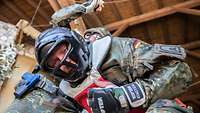 Ein Soldat schlägt vor einem Haus auf einen Soldaten mit Kopfschutz und Weste ein, während dieser in die Kamera schaut