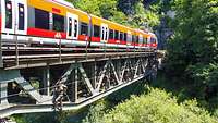 Soldatinnen und Soldaten des MN KdoOpFü überqueren kletternd eine Eisenbahnbrücke über der Donau, während dort ein Zug fährt.