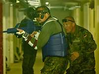 Nachttraining im Bunker in einem Kellergang ein gepolsterter Soldat mit Nachsichtbrille und Gewehrattrappe in Angriffshaltung, dahinter ein Trainer