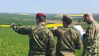 Drei Soldaten stehen auf einer Wiese, zwei zeigen in die Landschaft, einer hält eine Karte.