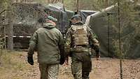 Zwei Soldaten gehen durch den Wald. Vor ihnen befinden sich mehrere große Zelte.