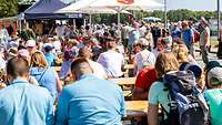 Viele Menschen sitzen auf Bänken an Tischen unter Sonnenschirmen