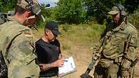 Zwei deutsche Soldaten und ein US-Soldat stehen beieinander. Der Amerikaner zeigt mit einem Stift auf einen Auswertebogen. 