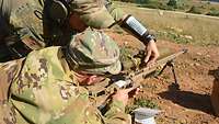 Ein US-Soldat liegt hinter einem Scharfschützengewehr G22A2. Ein deutscher Soldat hilft ihm.