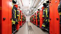 In einem langen schmalen hell erleuchteten Umkleideraum stehen rote Metallschränke in denen Feuerwehr-Uniformen hängen