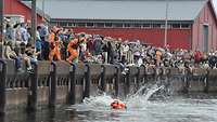 Zahlreiche Zuschauen schauen auf eine Person, welche im Hafenbecken in einem orangenem Rettungsanzug schwimmt.