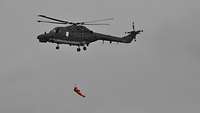Eine Person in einem orangenen Rettungsanzug hängt mit einem Seil an einem Hubschrauber.