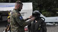 Ein Soldat in Flecktarn hilft einer kleineren Person beim Anlegen eines Pilotenhelmes.