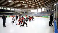 Besprechung mit dem Bundestrainer auf einem Eishockeyfeld knien Spielerinnen in Trikots und hören dem Trainer zu, der etwas an eine Tafel schreibt