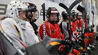 Auf einer Bank sitzen einige Eishockeyspielerinnen in Trikots (rot, schwarz und weiß) und Helm mit ihren Schlägern