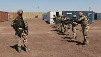 Ein Soldat steht vor einer Reihe von Soldaten mit Gewehren, im Hintergrund Container