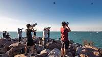 Mehrere Personen stehen am Strand und filmen in den Himmel