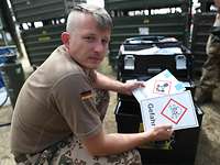 Warnschilder Ein Soldat hockt vor einem Transportkoffer und hält zwei Schilder mit der Aufschrift „Gefahr“ und „Achtung“ in den Händen.