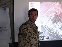 Taskforce-Leiter Ein Soldat zeigt mit einem Zeigestock auf eine Beamer-Projektion, die ein Luftbild von einem Gelände zeigt.