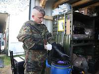 Ein Soldat im Schutzanzug steht vor einem Container mit Ausrüstung und zieht Latexhandschuhe an.