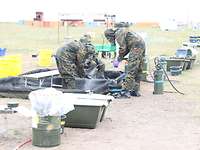 Soldaten in Schutzanzügen und mit ABC-Masken stehen in einer Wanne und besprühen sich.