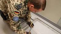 Ein Soldat kniet auf dem Boden und schraubt eine Rahmenhalterung vom Boden ab
