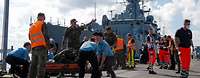 Zahlreiche Helfer auf einer Pier im Hintergrund eine Fregatte