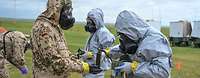 Mehrere Soldaten in ABC-Schutzkleidung stehen im Gelände und messen mittels Messgeräten die Kontamination ihrer Kleidung.