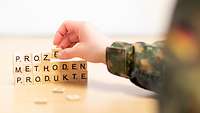 Scrabblesteine mit dem Schrisftzug "Methoden, Produkte und Prozesse werden von einem Soldaten übereinander gestapelt. 