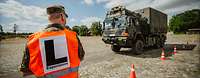 LKW mit Zuladung auf der Teststrecke Ein Soldat mit Warweste und Aufschrift „L–Experiment“ steht mit dem Rücken zum Betrachter vor einem LKW auf einer Teststrecke.