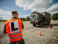 LKW mit Zuladung auf der Teststrecke Ein Soldat mit Warweste und Aufschrift „L–Experiment“ steht mit dem Rücken zum Betrachter vor einem LKW auf einer Teststrecke.