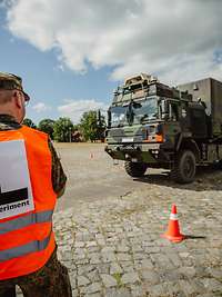 LKW mit Zuladung auf der Teststrecke Ein Soldat mit Warweste und Aufschrift „L–Experiment“ steht mit dem Rücken zum Betrachter vor einem LKW auf einer Teststrecke.