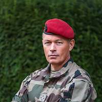 Ein General mit rotem Barett und verschränkten Armen schaut ernst in die Kamera. 