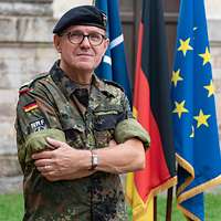 Ein General mit schwarzem Barett schaut in die Kamera, hinter ihm drei aufgestellte Flaggen.