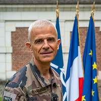 Ein General mit grauen kurzen Haaren schaut freundlich in die Kamera, hinter ihm die Nationalflagge.