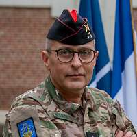 Ein General mit dunkelblauem Schiffchen vor Nationalflaggen hat die Arme verschränkt.