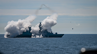 Ein graues Kriegsschiff in See, halb in zwei große Rauchwolken gehüllt.