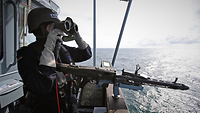 Ein Marinesoldat in blauer Arbeitsuniform mit Gefechtshelm hält ein Fernglas in beiden Händen.